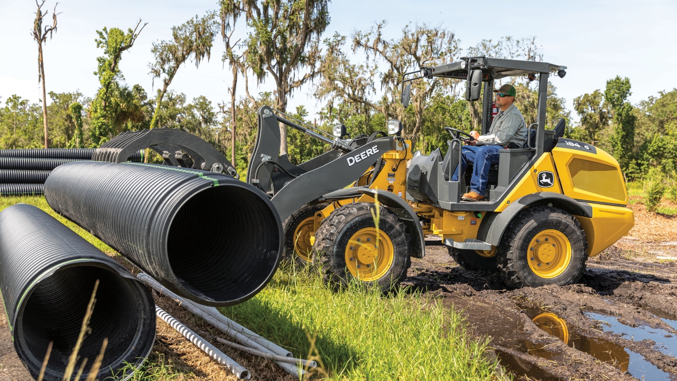 John Deere 184G Compact Wheel Loader with grapple attachment lifting a pipe.